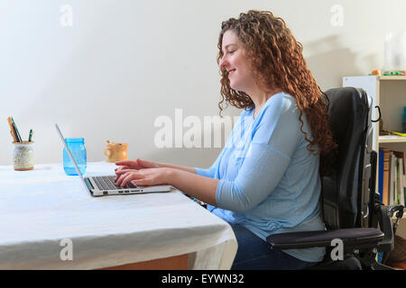 Donna con la distrofia muscolare in suo potere sedia usando il suo computer portatile nel suo ufficio Foto Stock