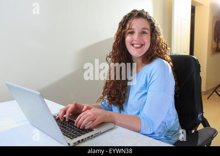 Donna con la distrofia muscolare in suo potere sedia usando il suo computer portatile nel suo ufficio Foto Stock