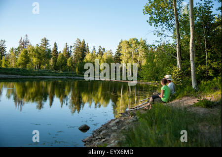 Una ragazza e il padre la pesca in una bella riflessione sul lago di montagna. L'acqua liscia riflette gli alberi in sera sun Foto Stock