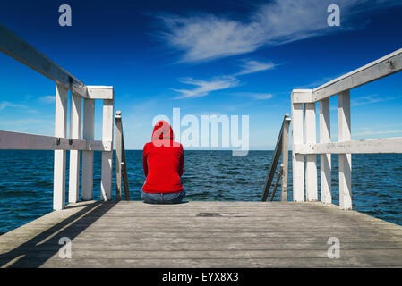 Da solo giovane donna in rosso Maglia con cappuccio seduta sul bordo del molo in legno guardando l'acqua - Disperazione, solitudine, alienazione C Foto Stock