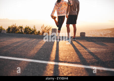 Ritagliato colpo di uomo e donna in piedi su una collina strada in mattina. Donna stretching la sua gamba. Paio di jogger guardando sunri Foto Stock
