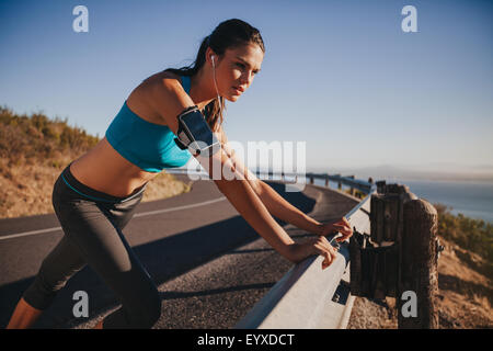 Giovane atleta femminile poggiando su autostrada guardrail guardando lontano. Donna runner fuori sulla strada di campagna prendendo una pausa dopo runni Foto Stock