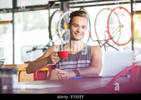 Uomo sorridente di bere il caffè al computer portatile in bike shop Foto Stock