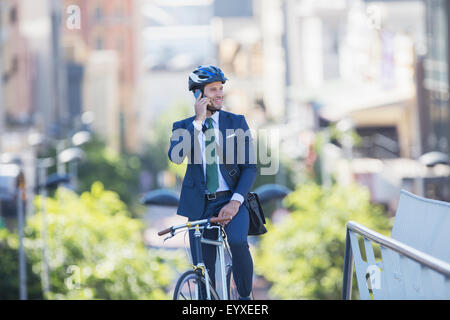 Imprenditore della tuta e casco seduto sulla bicicletta parlando al cellulare in città Foto Stock