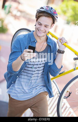 Giovane uomo con il casco che trasportano in bicicletta e i messaggi di testo sul telefono cellulare Foto Stock