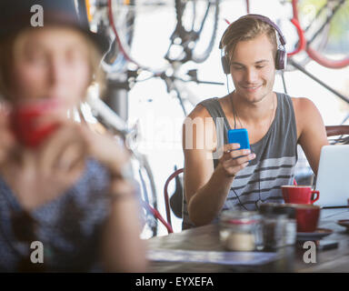 Giovane uomo con le cuffie e il computer portatile sms con il telefono cellulare in cafe Foto Stock