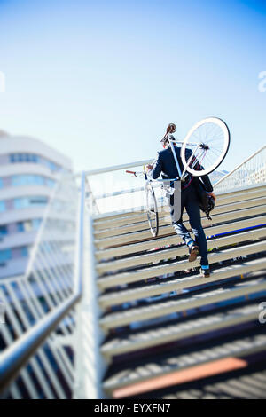 Imprenditore che porta bici fino urban scale sotto sunny blue sky Foto Stock
