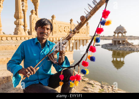 Uomo indiano holding strumento tradizionale vicino al monumento, Jaisalmer, Rajasthan, India Foto Stock