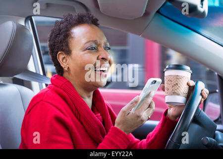 Nero donna tramite telefono cellulare e di bere il caffè mentre si guida auto Foto Stock