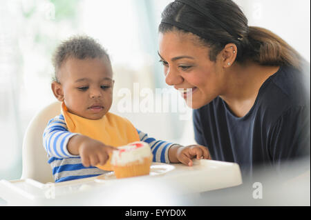 Razza mista madre dando baby figlio cupcake in sedia alta Foto Stock