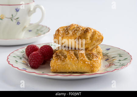 Tre pasticcini disposta accanto a un cluster di lamponi su un floreale piastra modellata con una corrispondente tazza e piattino dietro Foto Stock