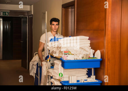 Giovane uomo spingendo un carrello pulizie laden con asciugamani puliti, servizio lavanderia e le apparecchiature di pulizia in un hotel come he services la camera Foto Stock