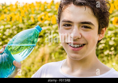 Eco-friendly living - Caucasian boy in un campo di girasoli è circa di bere acqua limpida da una bottiglia di plastica verde bluastro per dissetarsi in una calda e luminosa giornata estiva Foto Stock