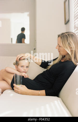 Donna con capelli lunghi biondi indossando un abito nero, seduto su un divano, tenendo in mano un bicchiere, giocando con sua figlia. Foto Stock