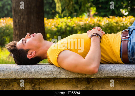 Attraente giovane uomo dorme sul banco in pietra all'aperto nel parco della città durante il giorno Foto Stock