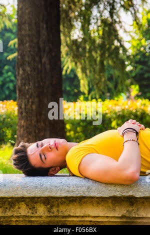 Attraente giovane uomo dorme sul banco in pietra all'aperto nel parco della città durante il giorno Foto Stock