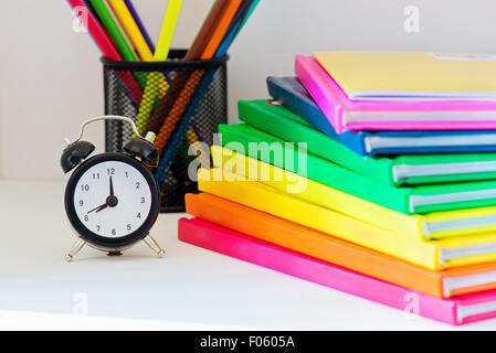 Concetto di istruzione. Nero orologio sveglia. Colorato libri in pila sulla luce color scaffale e matite a caso sul Foto Stock