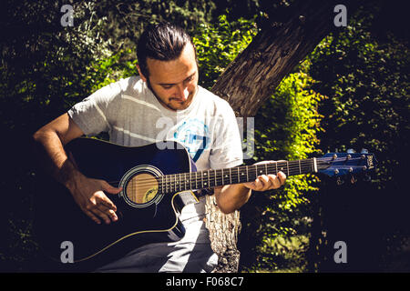 Lettore di chitarra a cantare in un parco seduto su una struttura ad albero Foto Stock