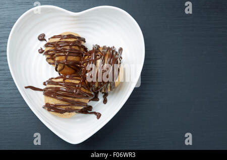 Chiudere fino ad alto angolo di visione dei tre gustosi dolci con salsa di cioccolato su un cuore bianco piastra sagomata, servita su una tabella di colore grigio con Foto Stock