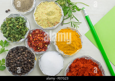 Varie spezie in ciotole lucido con una penna di colore verde e il libro verde per la scrittura di ricette su un bianco tavolo in legno Foto Stock