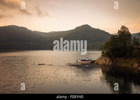 Piccola barca sul lago artificiale dietro la diga di Vidraru, al tramonto, Romania Foto Stock