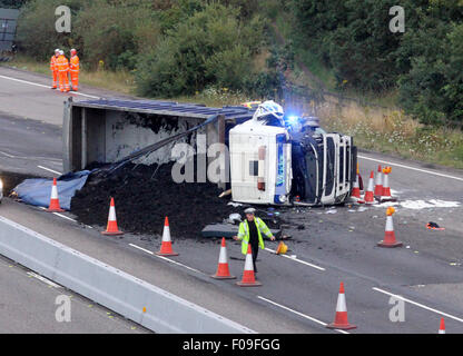 M3, Farnborough, Hampshire, Regno Unito. 10 Agosto, 2015. M3 vicino a Farnborough mostra GV della scena dopo un automezzo entra in collisione con una manutenzione autostrade veicolo parcheggiato sul disco spalla. Il carrello è stato dare protezione ai camion di recupero che stava recuperando un ripartiti automobilista. Il HGV dopo la alta velocità di collisione è venuto a poggiare sul suo lato dopo versando il suo carico su corsie uno e due del carrello principale modo. Due persone sono state portate in ospedale, uno da Air Ambulance la seconda da strada. Credito: Jason Kay/Alamy Live News Foto Stock