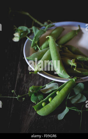 Cumulo di giovani piselli verdi intere e rotte con foglie e fiori in vintage piastra cena su un vecchio tavolo di legno. Rustico scuro styl Foto Stock