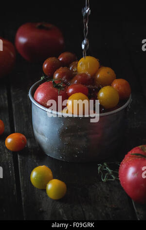 Versare acqua colorata a pomodori ciliegia in coppa in alluminio su un vecchio tavolo di legno. Scuro in stile rustico. La luce del giorno. Foto Stock