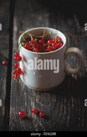 Vecchia coppa bianco pieno di fresche e mature di ribes rosso su un vecchio tavolo di legno. Scuro in stile rustico. La luce naturale del giorno. Foto Stock