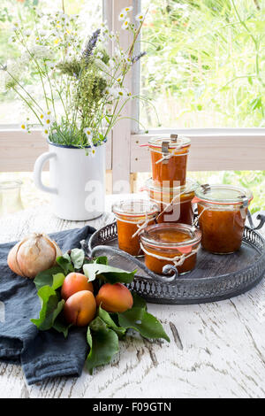 Chutney di albicocche vasetti sul vassoio vintage, con il grigio asciugatutto, bouquet di fiori e frutta fresca su bianco tavolo in legno Foto Stock