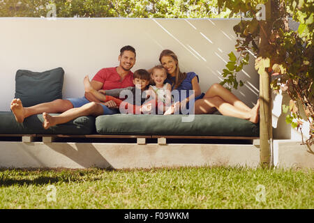 Ritratto di felice famiglia giovane seduto sul patio sorridente alla fotocamera. Coppia con bambini seduti sul divano nel loro cortile. Foto Stock