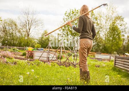 Donna matura, all'aperto, giardinaggio, portando rastrello, vista posteriore Foto Stock