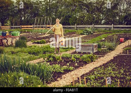 Donna matura, giardinaggio, portando rastrello e annaffiatoio Foto Stock