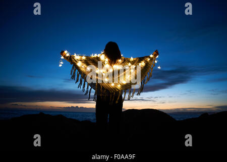 Vista posteriore della donna avvolta in scialle con stelle le luci fairy sulla spiaggia di notte Foto Stock