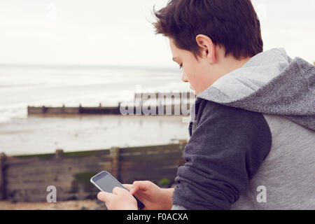 Ragazzo adolescente in riva al mare a scrivere messaggi su smartphone, Southend on Sea, Essex, Regno Unito Foto Stock