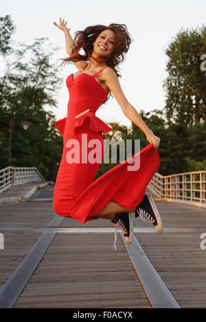 Ritratto di una giovane donna in un elegante vestito rosso e scarpe da baseball jumping in movimento Foto Stock