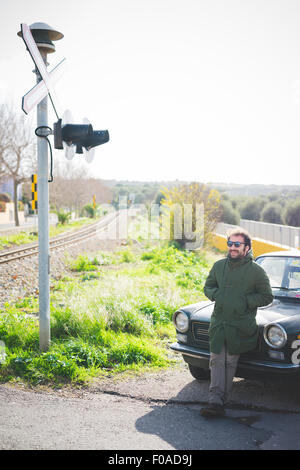 Metà uomo adulto appoggiata contro l'auto d'epoca a incrocio ferroviario Foto Stock