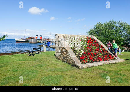 Il lungomare di Brodick sull'isola di Arran in Scozia con sollevati letto di fiori e di battelli a vapore Waverley presso il molo. Foto Stock