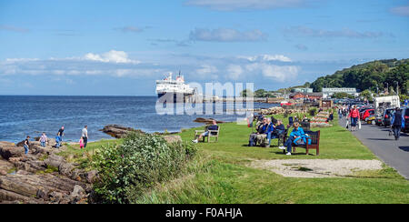 Il lungomare di Brodick sull'isola di Arran in Scozia con i turisti in corrispondenza di sedi godendo il sole e la Caledonian Isles traghetto per auto dietro. Foto Stock