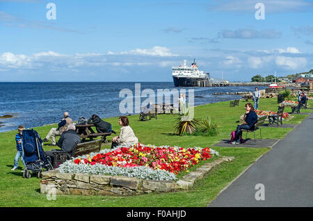 Il lungomare di Brodick sull'isola di Arran in Scozia con i turisti in corrispondenza di sedi godendo il sole e la Caledonian Isles traghetto per auto dietro. Foto Stock