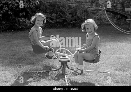 Due giovani ragazze che indossano pantaloncini pinafore giocando su un merry-go-round nel giardino. Foto Stock