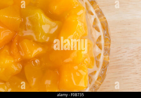 Top vista ravvicinata di una piccola ciotola di vetro di dadini di pesche in sciroppo pesante su una tavola di legno top illuminato con luce naturale. Foto Stock