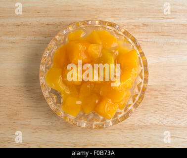 Vista dall'alto di una piccola ciotola di vetro di dadini di pesche in sciroppo pesante su una tavola di legno top illuminato con luce naturale. Foto Stock