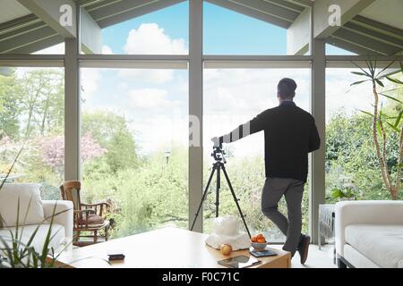 Vista posteriore dell'uomo appoggiata sul telescopio guardando fuori della finestra ammirando garden New Scenic 5 posti Foto Stock