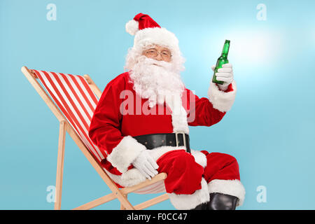 Santa Claus in possesso di una bottiglia di birra seduti su una sedia a sdraio e guardando la telecamera con cielo blu in background Foto Stock