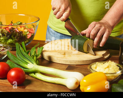 Insalata fresca ingredienti sul tavolo, uomo di mezza età taglia le zucchine - sfondo giallo Foto Stock