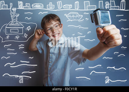 Un ragazzo di dieci anni di aspetto europeo con gli occhiali tenendo una telecamera nelle mani del sé su sfondo grigio Foto Stock