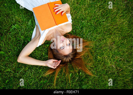 Una giovane donna si rilassa con un libro sdraiati su un prato verde nel parco. Giorno d'estate. Foto Stock