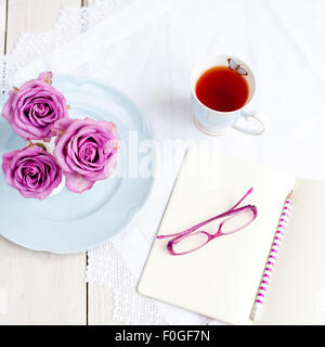 Tre rose rosa in vasi sulla piastra blu con la tazza di tè e aprire il notebook con gli occhiali e matita Foto Stock