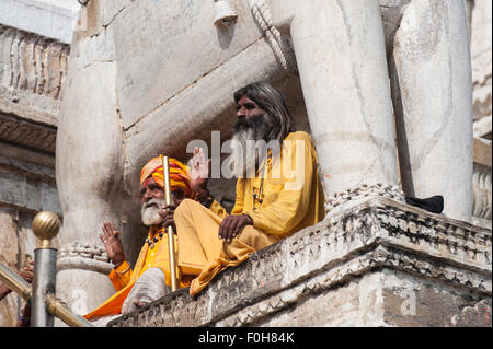 Udaipur, India. Sadhu indù santo uomini vestiti in abiti dello zafferano seduti sotto la statua di un elefante all'ingresso del xvii secolo Jagdish tempio con le loro mani sollevate nel messaggio di saluto. Foto Stock